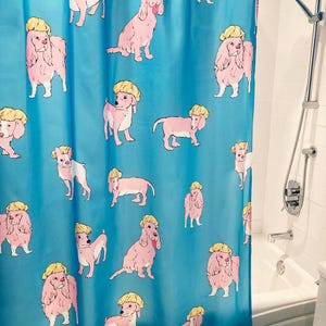 Op de afbeelding: Blauw douchegordijn met een herhaald patroon van roze honden met gele badmutsen. De honden staan in verschillende poses, wat een speels ontwerp creëert. Het gordijn hangt in een badkamer.