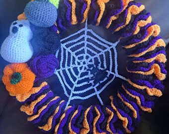 Könnte beinhalten: Ein gehäkelter Halloween-Kranz mit einem Spinnennetz-Design in der Mitte. Der Kranz ist mit gehäkelten Kürbissen, Geistern und einer grün-violetten Figur verziert. Der Kranz ist aus schwarzem, orangem und violettem Garn gefertigt.