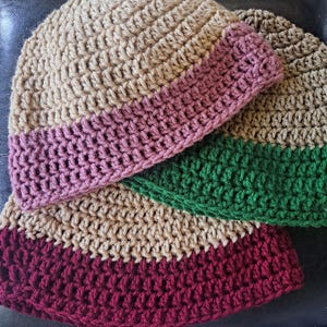 May include: Three handmade crochet beanies in shades of tan, burgundy, pink, and green. Each hat features a contrasting band at the brim. The hats are made with a textured stitch, perfect for cold weather.