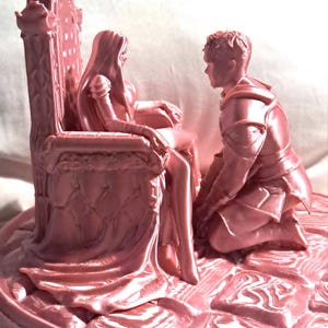 May include: A pink figurine of a medieval scene. A figure sits on an ornate throne, with another kneeling before them. The base is round. The overall design is a romantic, historical theme. The figurine is detailed and likely made of resin or a similar material.