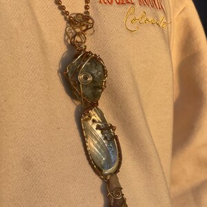 May include: A handcrafted pendant necklace featuring a large, irregularly shaped green stone, a pearlescent shell, and a purple crystal, all wrapped in copper wire. The necklace hangs from a delicate copper chain, creating a unique, earthy aesthetic.