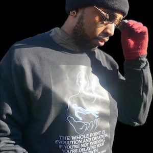May include: A black sweatshirt featuring a white graphic of a person in a hand, and the text "THE WHOLE POINT IS EVOLUTION AND CREATION." The person is wearing a black beanie, gold-rimmed glasses, and red gloves.