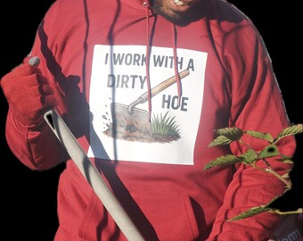 Op de afbeelding: Een rode hoodie met de tekst "I WORK WITH A DIRTY HOE" op de voorkant. De persoon op de foto draagt een zwarte muts, zonnebril en rode handschoenen, en houdt een tuinhark vast. De achtergrond is zwart.