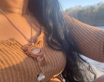 May include: A copper-colored pendant necklace with a large, oval-shaped stone is the focal point. The pendant is intricately wire-wrapped. The necklace is worn over a ribbed, tan-colored top. Long, dark hair frames the upper body.