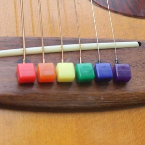 May include: A close-up of a guitar bridge with six strings. The strings are tied to colorful square beads in a rainbow pattern: red, orange, yellow, green, blue, and purple.