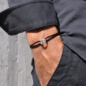 Könnte beinhalten: Ein schwarzes Kordelarmband mit einem silberfarbenen, knotenförmigen Mittelstück. Das Armband wird am Handgelenk getragen, mit einem dunkelgrauen Hemdsärmel im Hintergrund. Das Armband hat ein schlichtes, minimalistisches Design.