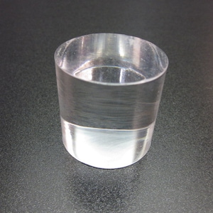 May include: A clear, cylindrical glass with a flat base. The glass is transparent, allowing light to pass through. The object is likely a small tumbler or a decorative piece.