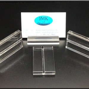 May include: Clear acrylic display stands in various shapes and sizes. The stands are arranged on a black surface, with a business card in the background that reads "JMK Displays". The stands are designed to showcase products.