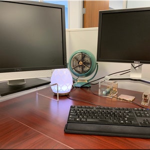 May include: A desk with two computer monitors, a keyboard, a white diffuser, a teal fan, and various office supplies. The desk is made of wood and has a light brown finish.