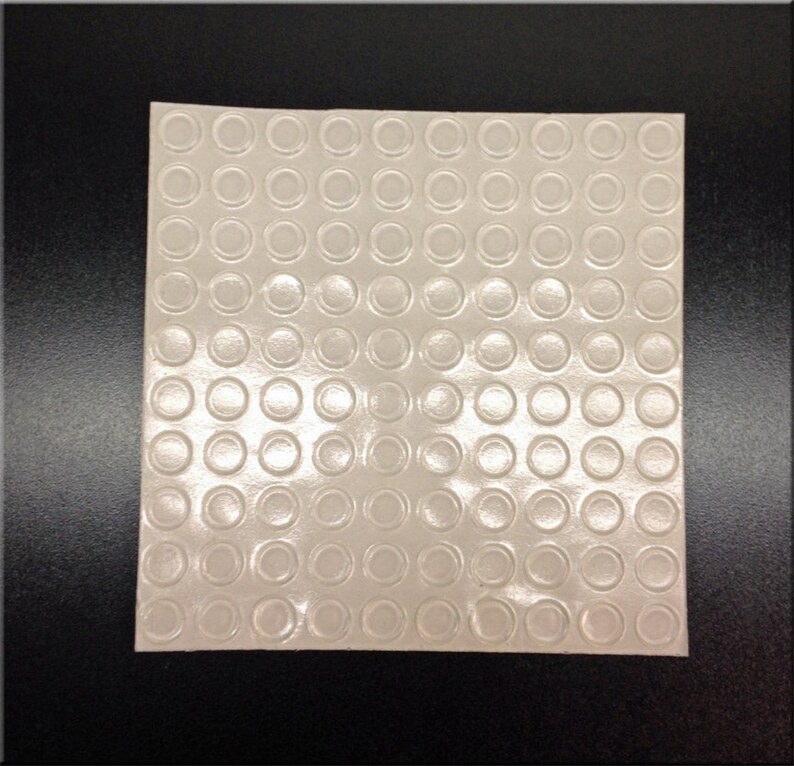 May include: A sheet of clear adhesive-backed rubber bumpers with a grid pattern of 10 rows and 10 columns. Each bumper is a small circle.
