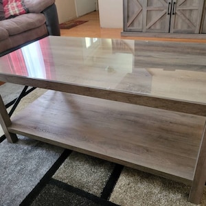 May include: A rectangular wooden coffee table with a glass top and a lower shelf. The table has a light brown wood finish and black metal accents.