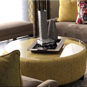 May include: A round, yellow, upholstered ottoman with a clear glass top. The ottoman has a geometric pattern and is decorated with three silver vases and a silver tray with a decorative box.