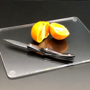 May include: A clear acrylic cutting board with rounded corners and four small, clear, circular feet. A small, black-handled knife and two halves of a peeled orange are on the board.