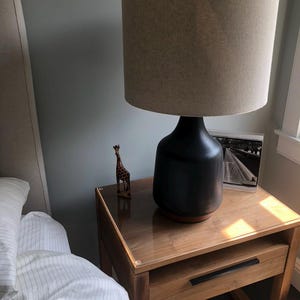 May include: A wooden nightstand with a drawer and a glass top. A black lamp with a beige lampshade sits on the nightstand. A small wooden giraffe figurine is also on the nightstand.