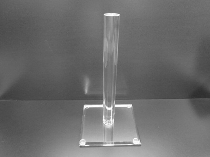 May include: Clear acrylic display stand with a square base and a tall, cylindrical post. The stand is transparent, allowing for a clear view of items placed on it. Ideal for showcasing jewelry, collectibles, or other small items.