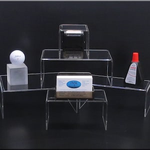 May include: Clear acrylic display stands of various sizes and shapes. A golf ball sits on a small cube. A bottle of red-capped adhesive and a business card holder are also displayed. The business card holder has the text "JMK Display Solutions".