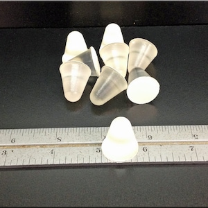 May include: A group of eight clear plastic cone-shaped bottle stoppers. The stoppers are approximately 2.5 cm in diameter and 2.5 cm in height. The stoppers are arranged in a random pattern on a black surface. A ruler is visible in the bottom of the image.
