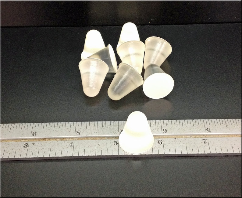May include: A group of eight clear plastic cone-shaped bottle stoppers. The stoppers are approximately 2.5 cm in diameter and 2.5 cm in height. The stoppers are arranged in a random pattern on a black surface. A ruler is visible in the bottom of the image.