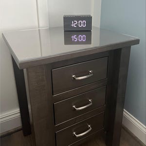 May include: A dark brown wooden nightstand with three drawers, each featuring a silver metal handle. The top surface is covered by a clear glass panel. A rectangular digital alarm clock with a black finish displays the time 12:00 in white light.
