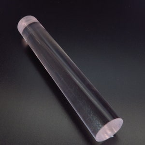 May include: A clear, cylindrical rolling pin made of transparent acrylic. The rolling pin is approximately 12 inches long and rests on a dark surface. The ends of the rolling pin are flat.