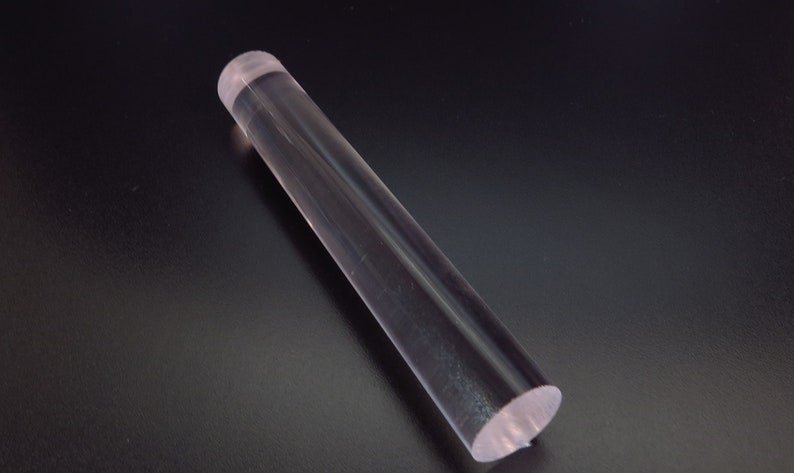 May include: A clear, cylindrical rolling pin made of transparent acrylic. The rolling pin is approximately 12 inches long and rests on a dark surface. The ends of the rolling pin are flat.