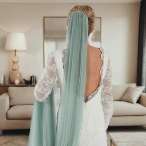 Peut inclure: Une femme portant une robe en dentelle blanche avec un dos nu et un long voile en tulle vert sauge. Le voile est drapé sur ses cheveux et descend le long de son dos.