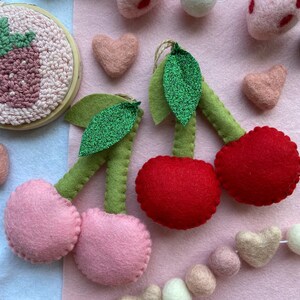 May include: A felt garland featuring pink and red cherries, green leaves, and white and pink felt balls. The garland is strung together with white thread.