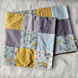 Könnte beinhalten: Zwei Patchwork-Babydecken mit verschiedenen Stoffen. Die Decken haben Quadrate mit gelben Punkten, Grau, lila Punkten und Hellblau mit Wal- und Wolkenmustern. Die Decken liegen auf einer weißen, flauschigen Oberfläche.