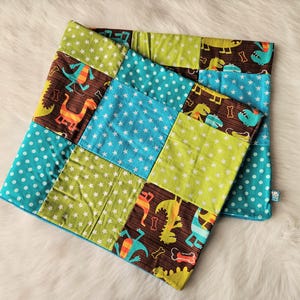 Könnte beinhalten: Eine Patchwork-Babydecke und ein passendes Kissen mit Dinosaurier-Motiv. Die Decke besteht aus Quadraten aus braunen, grünen und blauen Stoffen mit Dinosaurier- und Sternenmustern. Das Kissen hat ein ähnliches Design mit einem blauen Rand.