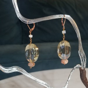 May include: A pair of dangle earrings with gold-colored leverback closures. Each earring features a large, faceted, light yellow-brown gemstone, a small, round, light pink gemstone, and a small white gemstone. The earrings are suspended from a gold-colored wire.