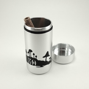 May include: A silver metal container with a black mushroom design and the word "KO". The container is open, revealing a cork inside. The lid is next to the container.
