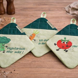May include: Three green and cream-colored pot holders with embroidered vegetable designs. One features a broccoli, another a carrot, and the last a tomato. Each has a green quilted top and a loop for hanging. Text is included on each pot holder.