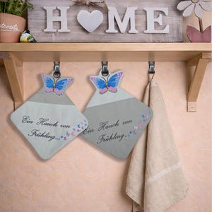 May include: Two diamond-shaped pot holders with butterfly designs and the text "Ein Hauch von Frühling..." hang below a shelf with a "HOME" sign. A beige towel hangs to the right. A small bird and potted plant are on the shelf.