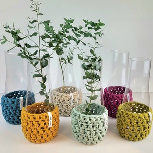 Könnte beinhalten: Eine Sammlung klarer Glasvasen mit gewebten, farbigen Hüllen. Die Hüllen sind in den Farbtönen Türkis, Gelb, Beige, Mintgrün, Lila und Olivgrün gehalten. Jede Vase enthält einen Zweig grünes Laub. Die Hüllen haben ein Etikett mit der Aufschrift "happy day".