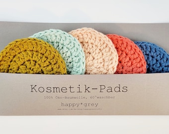 May include: A set of five colorful, crocheted cosmetic pads in a gray cardboard holder. The pads are in shades of yellow, mint green, peach, coral, and blue. Text on the holder reads "Kosmetik-Pads" and "happy*grey".