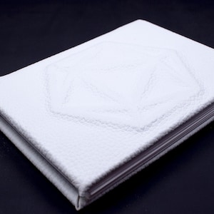 May include: A white leather-bound journal with a geometric design embossed on the cover. The journal has a blue ribbon bookmark.