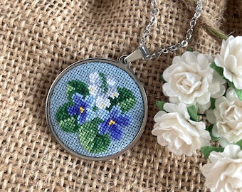Puede incluir: Un collar de cadena de plata con un colgante redondo que presenta un diseño de punto de cruz de flores moradas y blancas sobre un fondo azul.