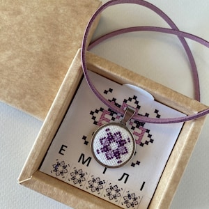 May include: A purple and white cross-stitch design on a round pendant necklace. The necklace is in a small box with the name "EMILI" printed on the front.