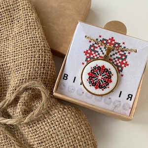May include: A gold necklace with a round pendant featuring a red and black cross-stitch design. The pendant is in a small box with the word "BI" on the front.