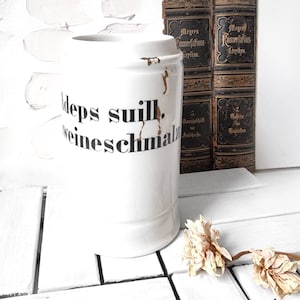 以下が含まれることがあります： 「Adeps suill. Weineschmalz」と書かれた黒い文字が入った白いセラミック容器。容器には蓋とハンドルが付いています。背景には、金色の文字とドライフラワーが施されたアンティークの本が含まれています。