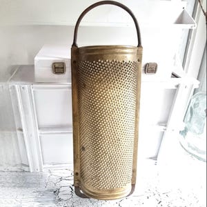 Könnte beinhalten: Ein Vintage-Käsehobel aus Messing mit einem perforierten zylindrischen Körper und einem gebogenen Griff. Der Hobel ist rechteckig mit abgerundetem Boden und zum Aufhängen konzipiert. Das Messing hat eine warme, gealterte Patina.