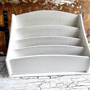 Può includere: Un organizer da scrivania in legno bianco con quattro scomparti curvi. L'organizer ha un design semplice e funzionale ed è verniciato con una finitura bianca opaca. La venatura del legno è visibile, conferendogli un aspetto rustico.