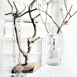 以下が含まれることがあります： 銀色のチェーンが流木で作られた彫刻からぶら下がっています。彫刻は木製の台座の上に置かれています。背景は白で、白い棚と綿の枝が入った透明なガラスの花瓶があります。