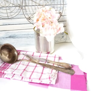 May include: A silver ladle and two silver forks on a pink and white checked kitchen towel. A can with a pink flower arrangement is in the background.