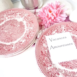 May include: Two pink and white floral patterned plates with a white card on top of one of the plates. The card has the text "Vacances Amoureuses" written on it.