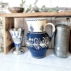 May include: A wooden shelf with three items: a blue and white ceramic jug with a floral design, a silver measuring cup with the text "Dr. Oetker Messbecher Measuring Cup", and a silver metal container with a handle.