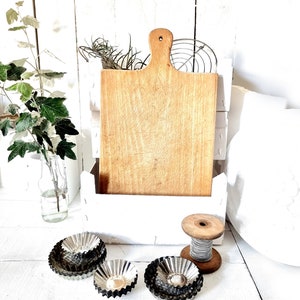 Könnte beinhalten: Ein weißes Holzregal mit einem Holzbrett zum Schneiden, einer Garnrolle und vier silbernen Cupcake-Förmchen. Das Regal ist mit einem weißen Blumendesign verziert.