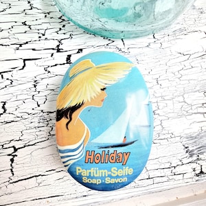 Può includere: Sapone ovale con un design vintage raffigurante una donna con un cappello da sole, una barca a vela e la parola "Holiday". Il sapone ha uno sfondo blu con le parole "Parfum-Seife Soap Savon."