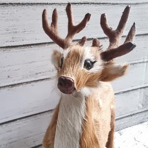 Op de afbeelding: Een handgemaakt hertenbeeldje met een pluche, getextureerde vacht van bruin en bruin bont. Het hert heeft grote, donkere ogen, een donkerbruine neus en gedetailleerde geweien. Het beeldje is een decoratief item, mogelijk voor feestdagen of seizoensgebonden weergave.
