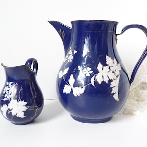 May include: Two blue enamel jugs with white floral designs. The larger jug has a handle and a spout. The smaller jug has a handle.